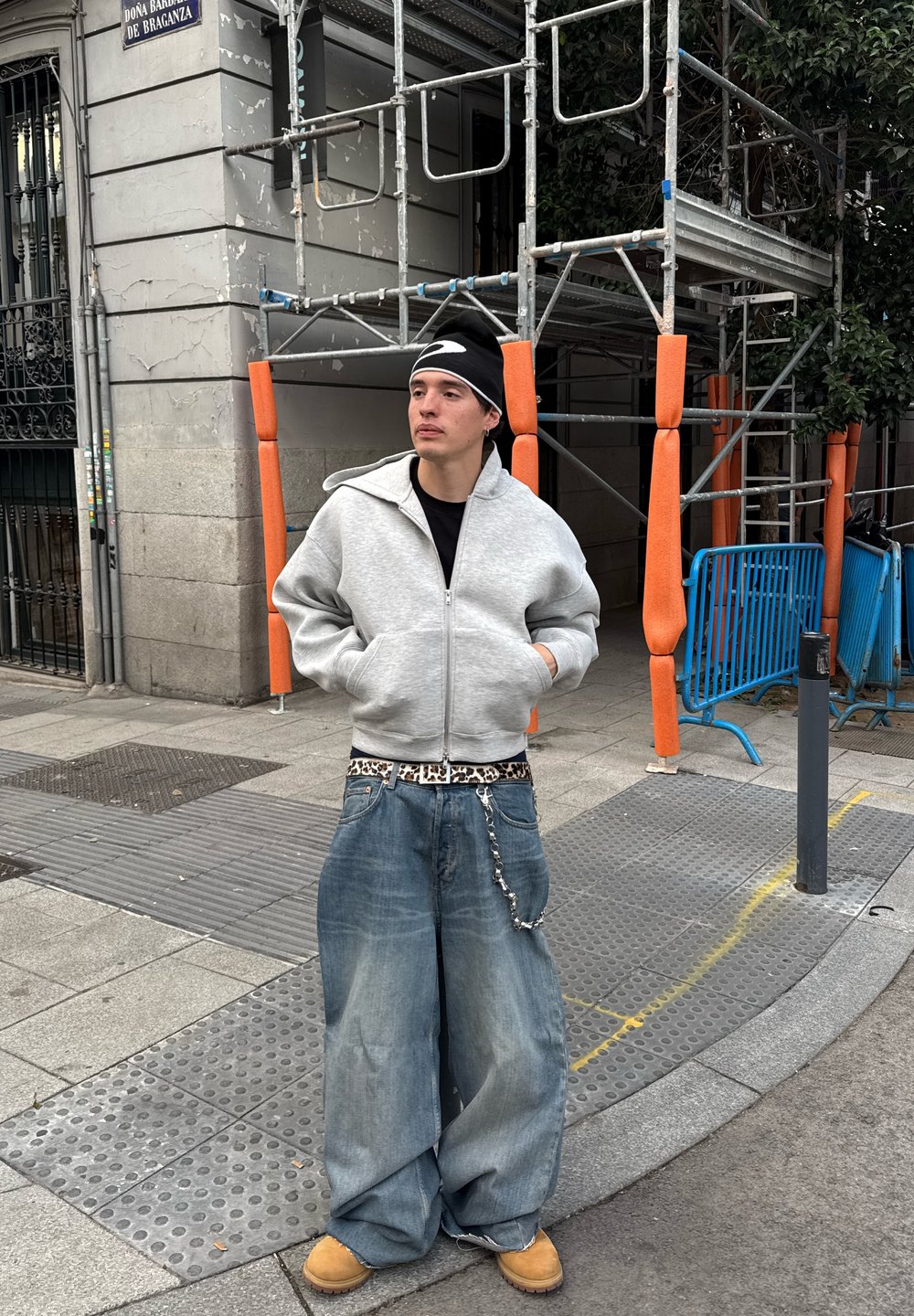 Pelēks kapucis ar rāvējslēdzēju, brīvi zili džinsus, dzīvnieku raksta josta, ikdienišķi apavi, melns beanie. Pilsētas vide ar būvscenēm un oranžiem akcentiem.