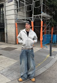 Pelēks kapucis ar rāvējslēdzēju, brīvi zili džinsus, dzīvnieku raksta josta, ikdienišķi apavi, melns beanie. Pilsētas vide ar būvscenēm un oranžiem akcentiem.