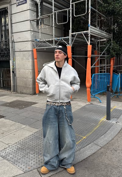 Sudadera gris con cremallera, pantalones de mezclilla azules holgados, cinturón de estampado animal, calzado casual, gorro negro. Entorno urbano con andamios y acentos naranjas.