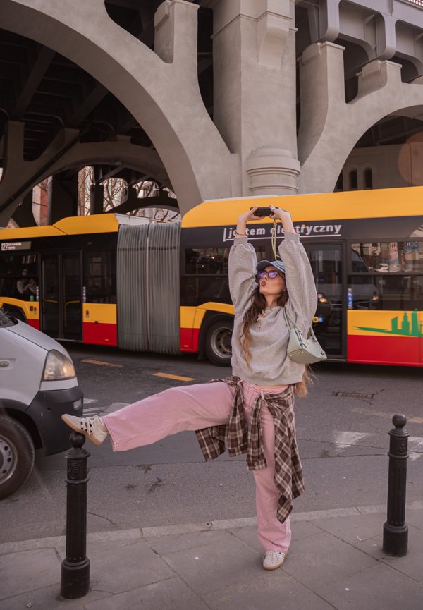 Fiatal nő rózsaszín nadrágban és szürke pulóverben egy lábon pózol a városi járdán, egy sárga és piros busz mellett, betonhíd alatt.