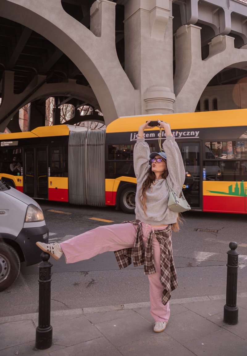 Mladá žena v ružových nohaviciach a šedej mikine pózuje na jednej nohe na mestskom chodníku pri žltom a červenom autobuse pod betónovým mostom.