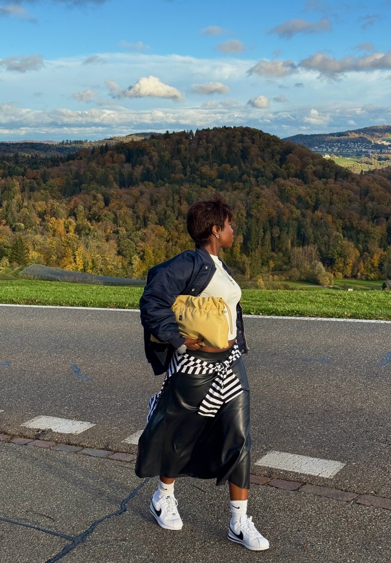 Svart läderkjol lager på ett randigt mönster, gul väska, marinblå jacka och vita sneakers. Utomhusmiljö med gröna kullar och molnig himmel.