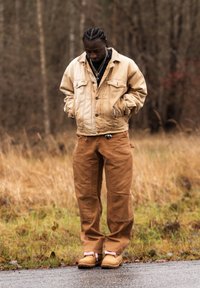 Homme aux cheveux tressés portant une veste beige superposée, un pantalon marron et des bottes, debout sur une route mouillée avec de l'herbe sèche et des arbres en arrière-plan.