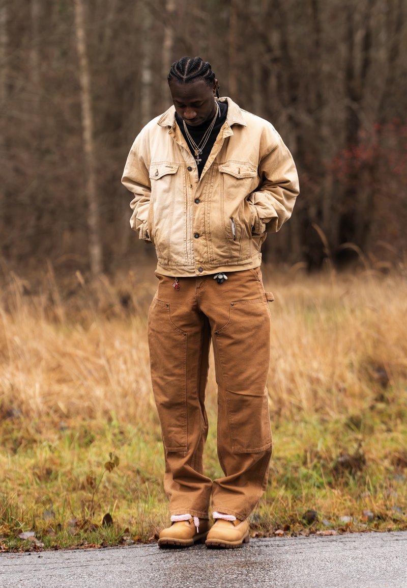 Muž s vrkočmi, ktorý má na sebe vrstvový svetlohnedý kabát, hnedé nohavice a čižmy, stojí na mokrej ceste s suchou trávou a stromami v pozadí.