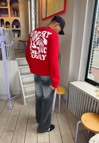 Suéter de punto rojo con el texto blanco "LO SIENTO, C HOY" en la parte de atrás, combinado con jeans de pierna ancha grises y zapatos negros sin cordones.