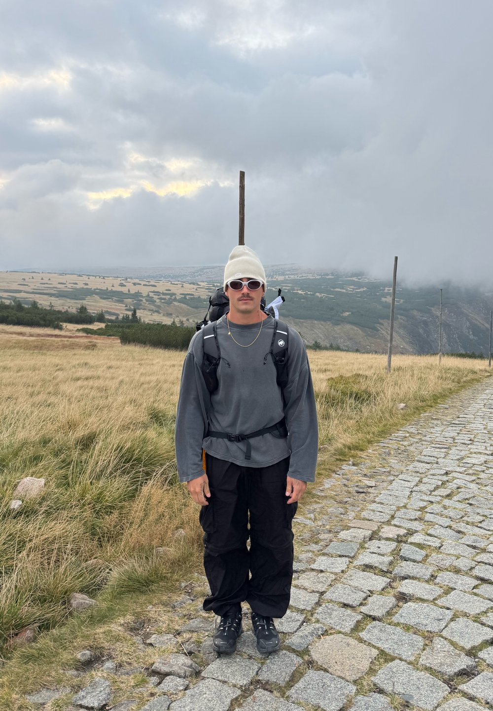 Čovjek u bijeloj kapi, sunčanim naočalama, sivom trenirci, crnim hlačama i planinarskim čizmama stoji na stazi od kaldrme u travnatom planinskom području.