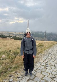 Čovjek u bijeloj kapi, sunčanim naočalama, sivom trenirci, crnim hlačama i planinarskim čizmama stoji na stazi od kaldrme u travnatom planinskom području.