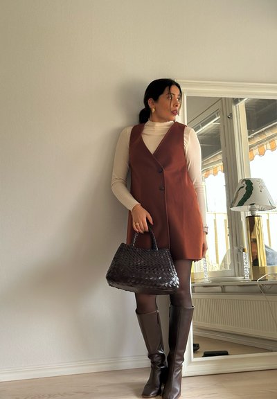 Vestido marrón sin mangas con dos botones, usado sobre un cuello de tortuga color crema. Un bolso negro de tejido y botas marrones hasta la rodilla completan el conjunto.