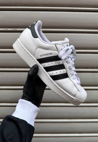 Χέρι με μαύρο γάντι κρατά λευκό αθλητικό παπούτσι Adidas Superstar με μαύρες ρίγες πάνω σε οριζόντιο μπεζ μεταλλικό φόντο.