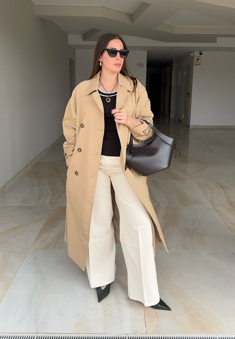 Femme portant un trench-coat beige, un haut noir, un pantalon large crème, des chaussures noires à bout pointu, des lunettes de soleil et un sac à main noir dans un couloir.