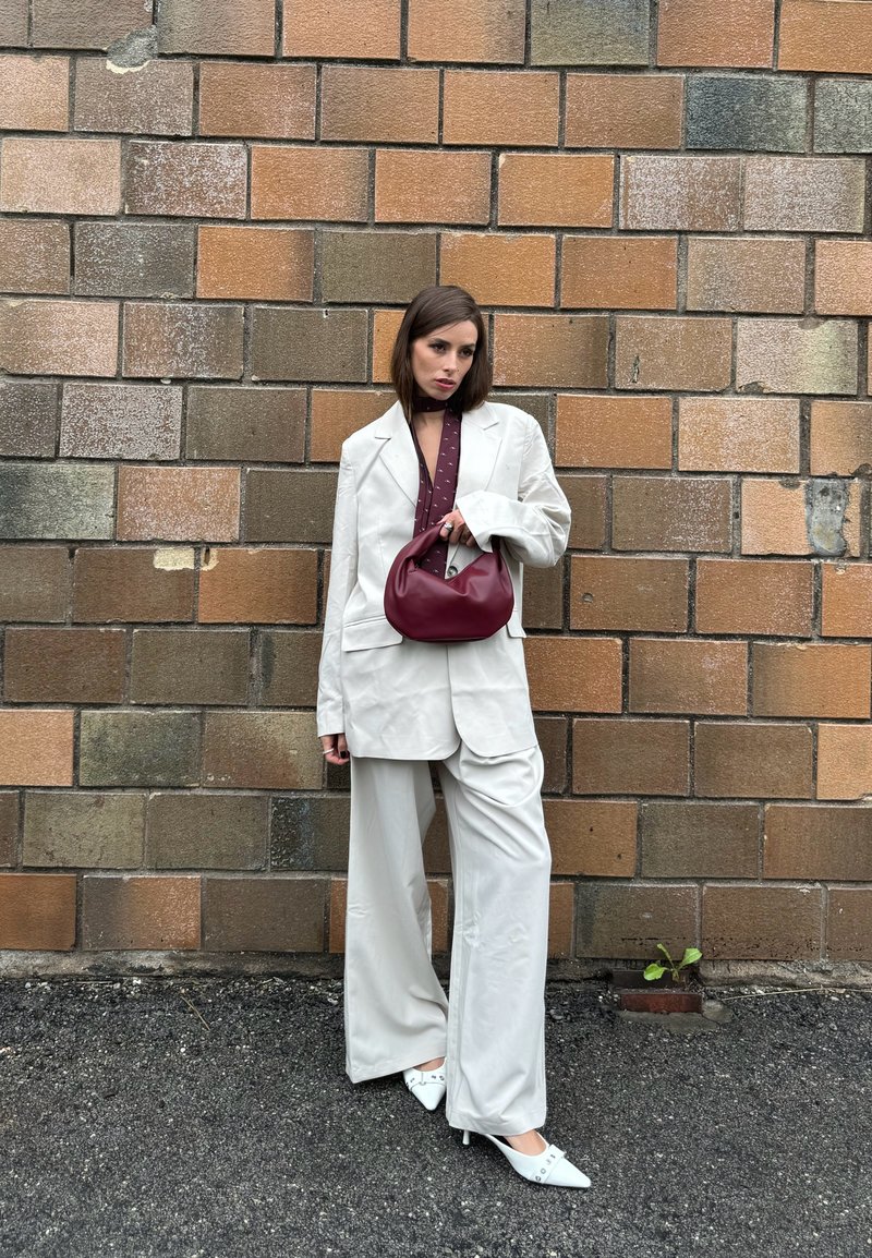 Blazer oversized creme com calças largas combinadas, acompanhados de uma bolsa em tons de maroon e sapatos de salto brancos de bico fino. Parede texturizada ao fundo.