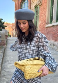 Szürke érzetű beret, áttetsző kockás blúz fekete-fehér színben, valamint egy sárga, háromszög alakú, font clutch táska. A texturált és rétegezett részletek láthatók.