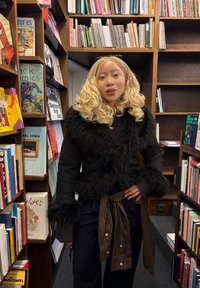 Casaco de pelo sintético preto com gola e punhos largos, usado sobre uma blusa preta. Gravata castanha com padrão xadrez na cintura, num espaço repleto de livros.