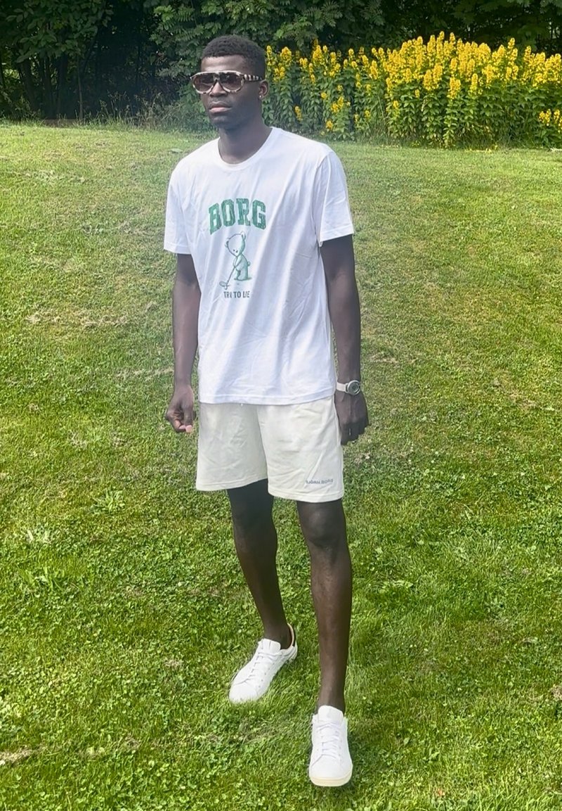 Homme portant des lunettes de soleil, un t-shirt blanc avec une impression verte, un short blanc et des baskets, debout sur de l’herbe près de fleurs jaunes.