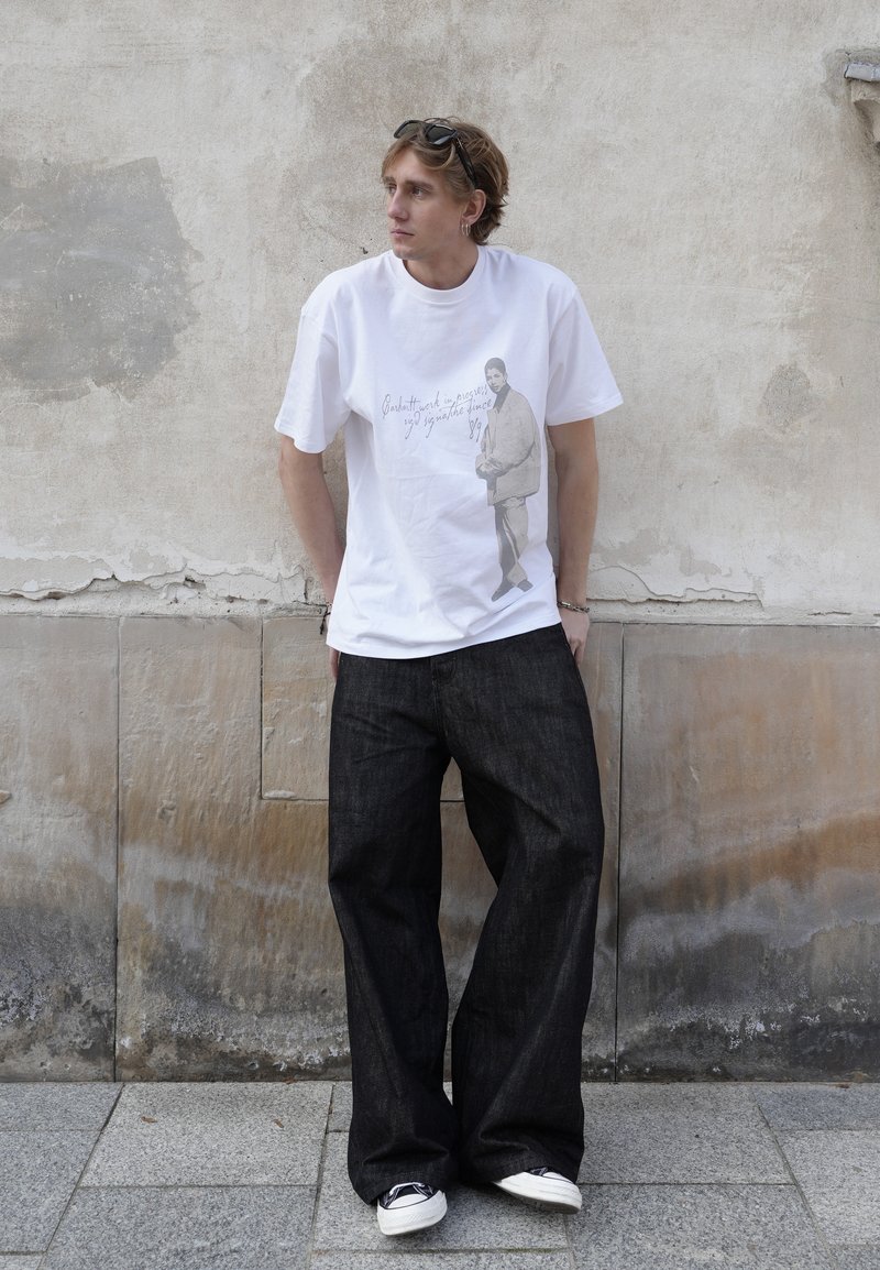 Un jeune homme portant un T-shirt blanc à motif graphique, un jean noir ample et des baskets blanches pose devant un mur de pierre patiné.