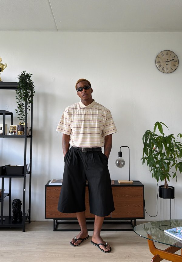 Homme portant des lunettes de soleil, un polo rayé, un short large noir et des tongs se tient dans une pièce moderne avec des plantes et des étagères.