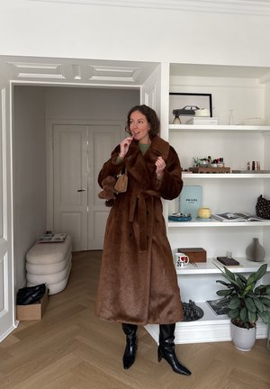 Mujer con un abrigo largo de piel marrón, botas negras y suéter verde, sosteniendo un bolso marrón, de pie en una habitación moderna con estanterías y plantas.