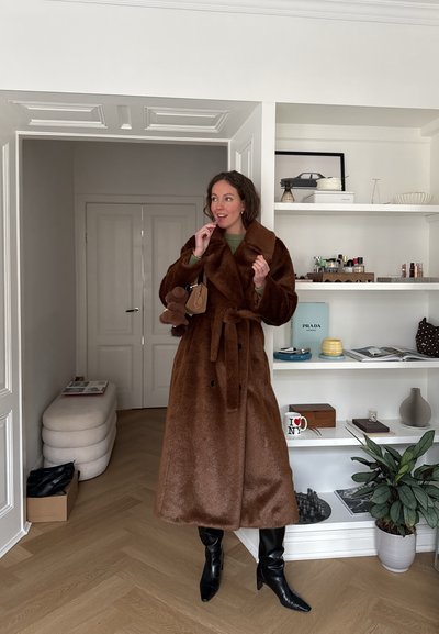 Mujer con un abrigo largo de piel marrón, botas negras y suéter verde, sosteniendo un bolso marrón, de pie en una habitación moderna con estanterías y plantas.