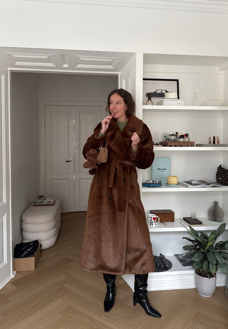 Femme portant un long manteau en fourrure marron, des bottes noires et un pull vert, tenant un sac à main marron, debout dans une pièce moderne avec des étagères et des plantes.