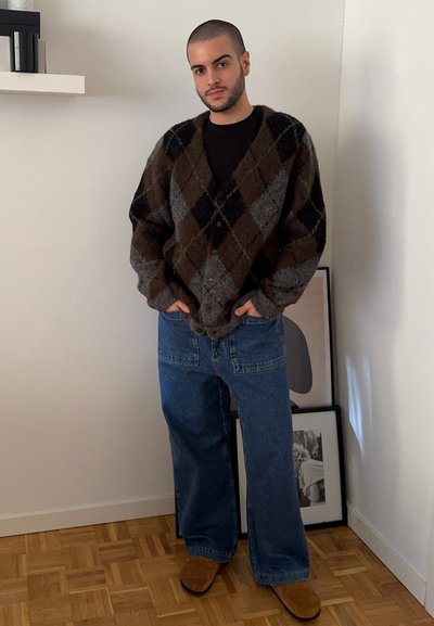 Cardigan de punto a cuadros en marrón y gris, de corte oversized, con una camiseta negra debajo, combinado con jeans de mezclilla azul holgados y zapatos marrones de deslizar.