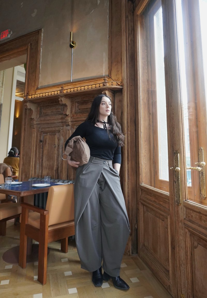 Jeune femme en haut noir et large pantalon gris debout près d'une grande fenêtre en bois tenant un sac marron à l'intérieur d'un café.