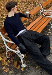 Persona seduta su una panchina di legno che indossa un maglione blu navy a trecce, pantaloni neri e guanti neri, tenendo in mano una tazza di caffè.