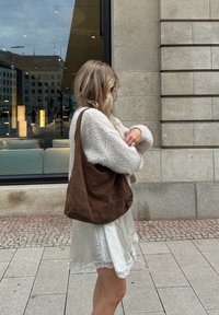 Ruda zomša petneška suknelė su minkštu, laisvalaikio dizainu. Modelis vilki kreminės spalvos megztinį ir baltą suknelę su nėriniuotais kraštais, stovi lauke.