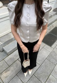 Blouse gris clair à manches bouffantes avec une fermeture à six boutons, associée à un pantalon noir à jambes larges. Tient un petit sac à main métallique texturé.