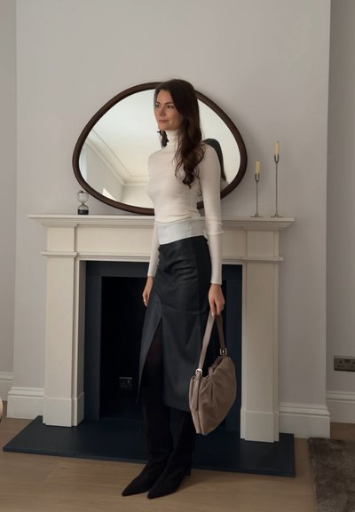 Mujer con suéter de cuello alto blanco, falda de cuero negra con abertura frontal, botas negras, sosteniendo un bolso beige, de pie junto a una repisa con espejo y velas.
