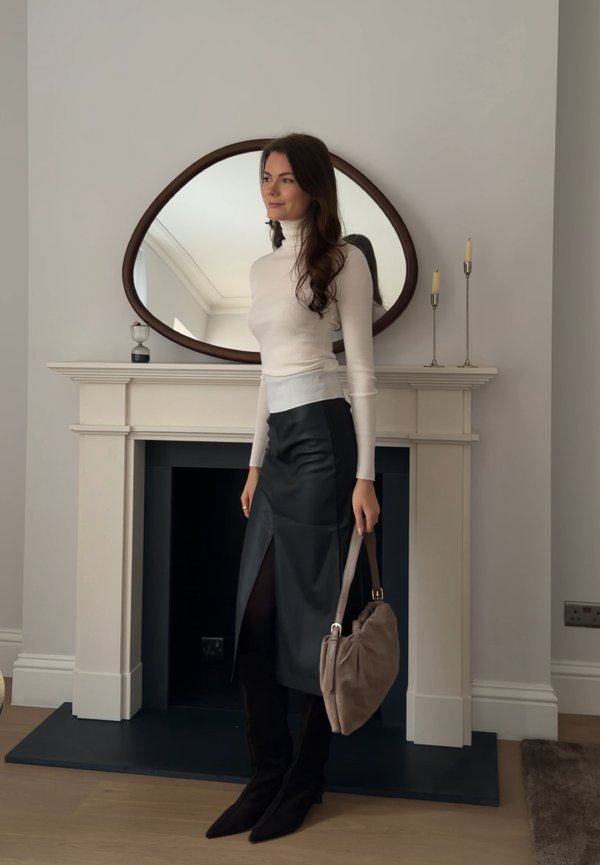 Ženska v belem puloverju z visokim ovratnikom, črni usnjeni krili s sprednjim razporkom, črnih škornjih, ki drži bež torbico, stoji ob kaminski polici z ogledalom in svečami.