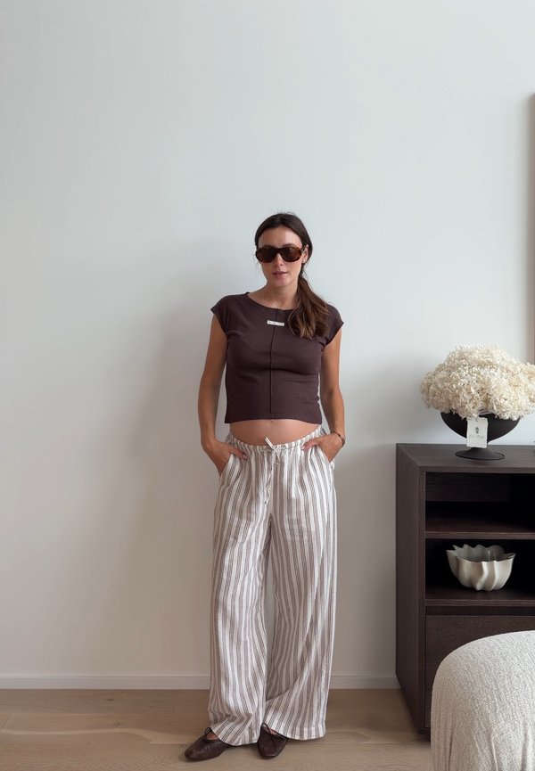 Mujer con gafas de sol, una camisa oscura recortada y pantalones sueltos a rayas, de pie con las manos en los bolsillos junto a un armario oscuro con flores blancas.