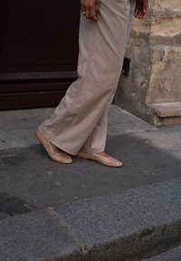 Beige flade sko med spids tå og prikkede hvide accenter, parret med lyse beige bukser med bred ben, på et mørkegrå pavement.