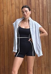 Čierny jednorazový overal s bielym lemovaním, doplnený svetlošedým, oversized mikinou prehodenou cez plecia. Hladká tkanina, priliehavý strih.