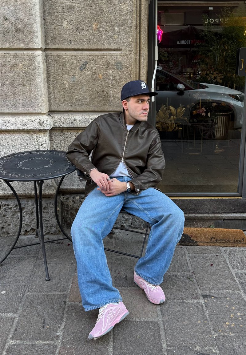 Homme en veste marron, casquette bleue, jean large et baskets roses assis sur une chaise en métal noir à côté d'une table ronde devant un bâtiment en pierre.