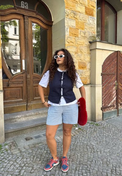 Chaleco azul marino sobre camiseta blanca, pantalones cortos a rayas y bolso de piel sintética roja. Gafas de sol blancas, zapatillas deportivas multicolor. Fondo de edificio de piedra.
