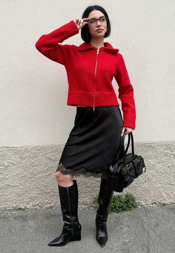 Kobieta o czarnych włosach obciętych na boba i w okularach, ubrana w czerwony sweter z zamkiem, czarną spódnicę z koronkowym wykończeniem, czarne kozaki za kolano, trzymająca czarną torebkę.