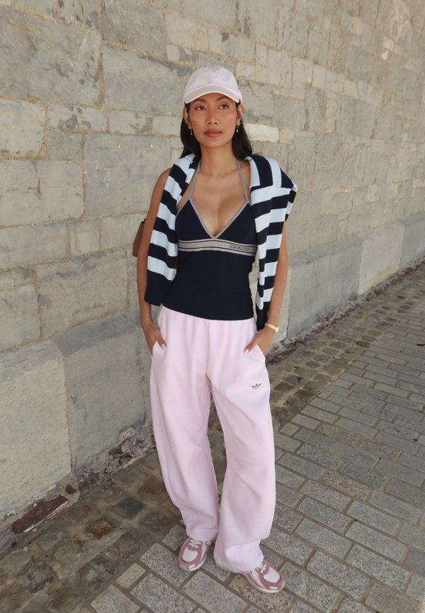 Jeune femme portant une casquette rose, un débardeur noir, un pull rayé sur les épaules, un pantalon rose ample et des baskets se tient sur un trottoir en pierre.