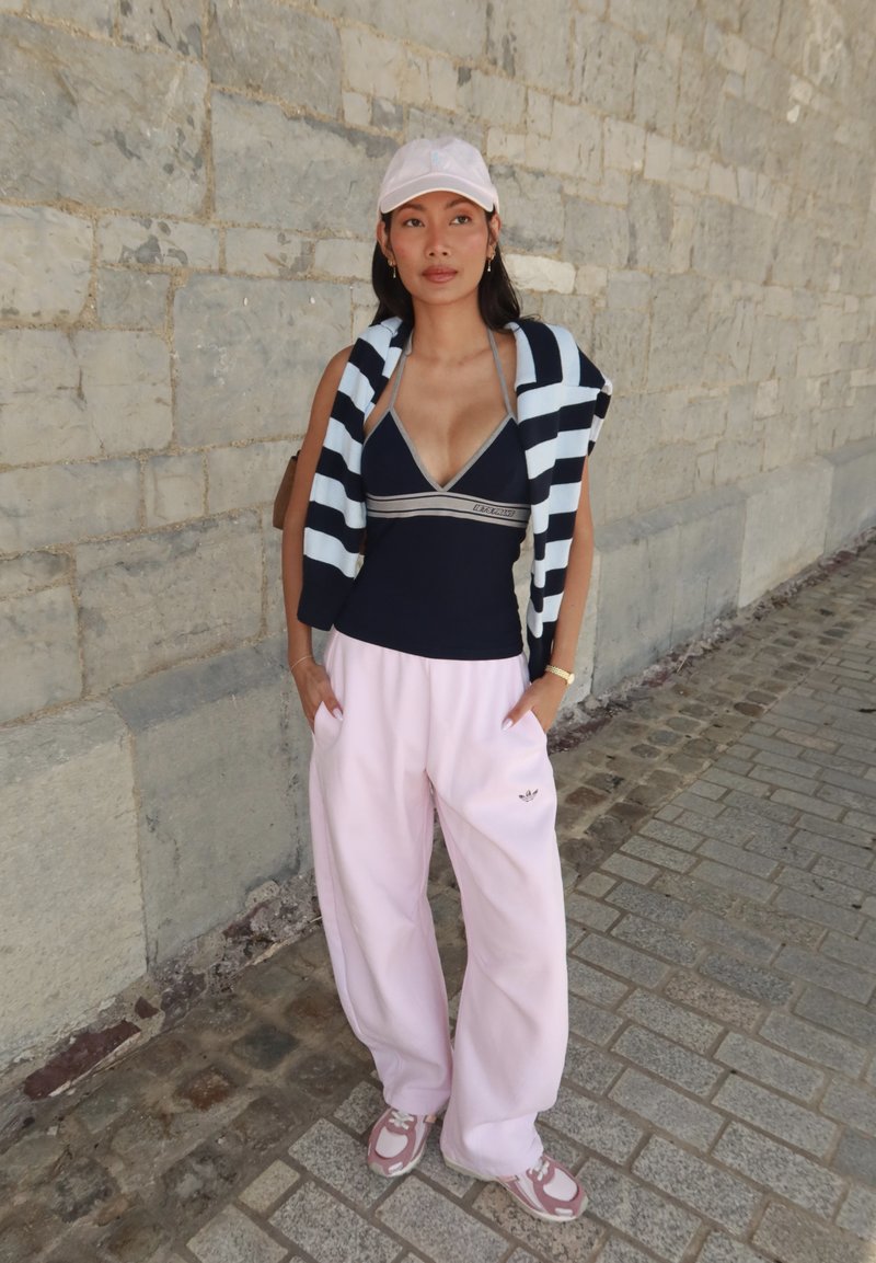 Giovane donna con cappellino rosa, canottiera nera, maglione a righe sulle spalle, pantaloni rosa larghi e scarpe da tennis, in piedi su un marciapiede di pietra.