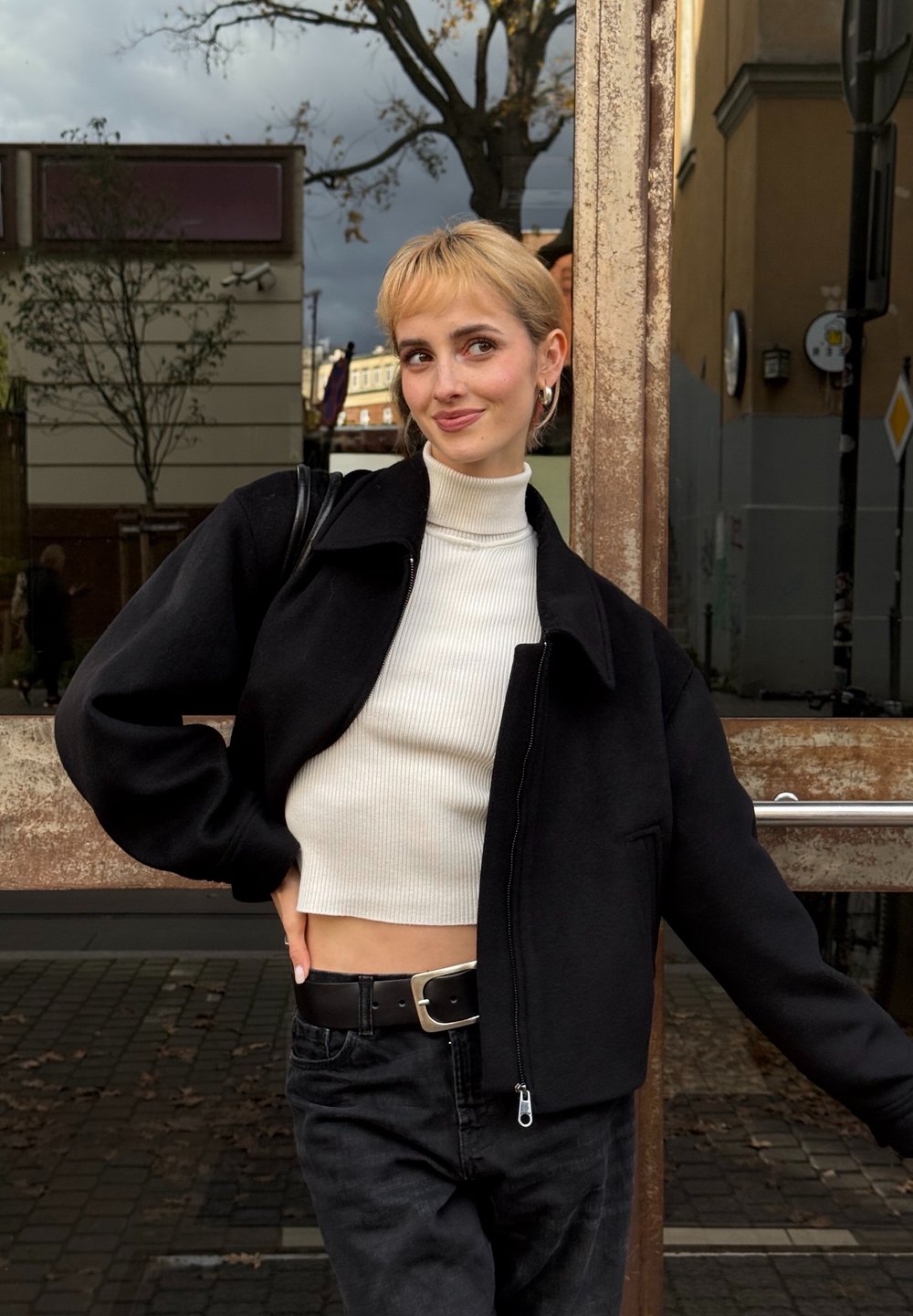 Junge Person mit kurzen blonden Haaren, die einen weißen, bauchfreien Rollkragenpullover und eine schwarze Jacke trägt und sich an einem rostigen Fensterrahmen im Freien anlehnt.