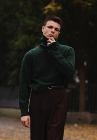 Maglione verde a collo alto in un tessuto testurizzato, abbinato a pantaloni scuri marroni su misura. Gli accessori in evidenza includono una catena d'argento e un anello.