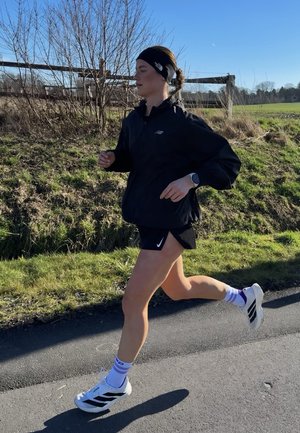 Mujer corriendo al aire libre por un camino pavimentado, vistiendo una chaqueta negra, pantalones cortos, diadema, calcetines blancos y zapatillas blancas, con césped y una cerca de madera cerca.
