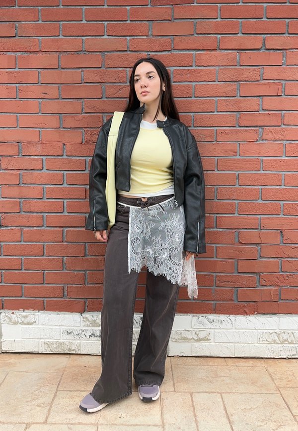 Γυναίκα που στέκεται μπροστά σε κόκκινο τοίχο από τούβλα, φοράει μαύρο δερμάτινο μπουφάν, κίτρινο πουκάμισο, καφέ φαρδιά παντελόνια, δαντελένιο φουλάρι και κρατάει μια κίτρινη τσάντα ώμου.