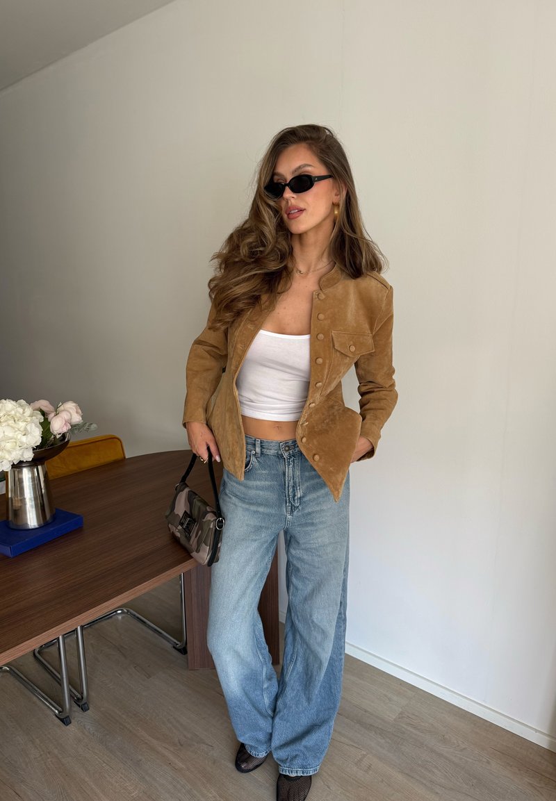 Femme aux longs cheveux ondulés portant des lunettes de soleil noires, une veste en daim marron, un crop top blanc, un jean à jambes larges, tenant un petit sac à main à l'intérieur près d'une table.