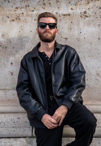 Giubbotto bomber di pelle nera con polsini a costine, indossato sopra una polo nera. Capelli acconciati e occhiali da sole indossati. Sfondo in pietra sobria.