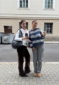 Zwei Frauen stehen neben einem Spiegel. Die eine trägt ein weißes Shirt und dunkle, braune Schlaghosen, die andere einen blau-gestreiften Cardigan und graue Jeans.