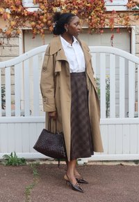 Beżowy trencz na białej koszuli, brązowa kraciasta spódnica, ciemna pleciona torebka oraz brązowe szpilki z ostro zakończonym noskiem. Plener z białą bramą.