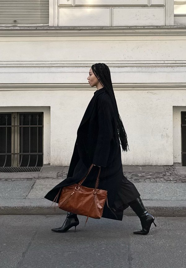 Woman with long braided hair walking along a city sidewalk wearing a black coat and boots, carrying a large brown leather handbag.