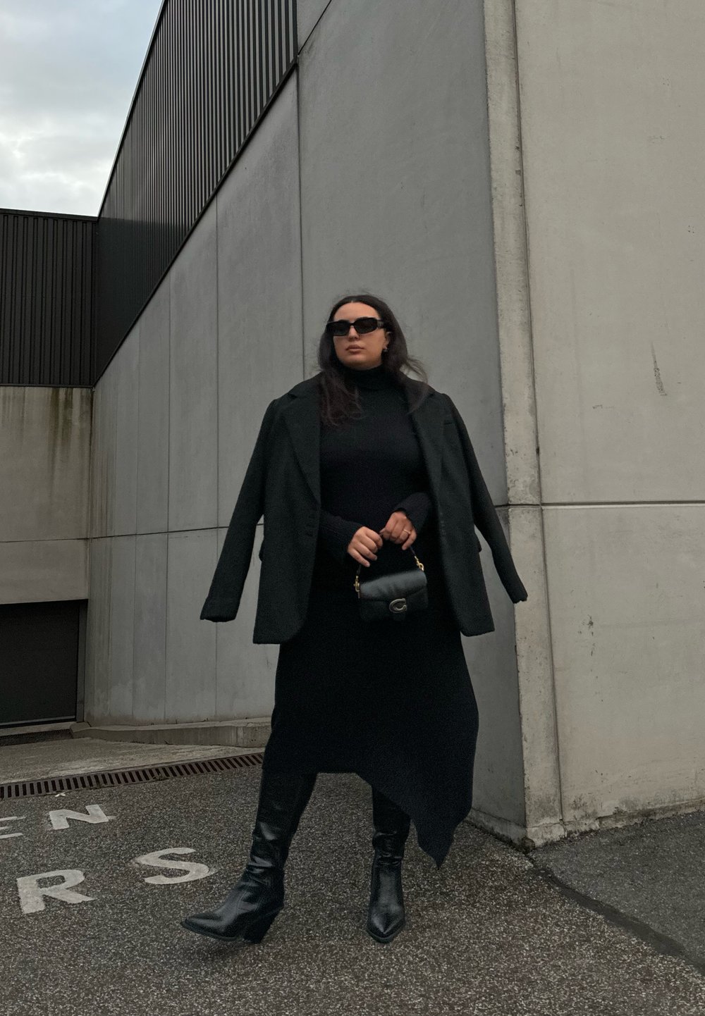 Črno oversized blazer nad prilegajočim črnim puloverjem s turtleneck ovratnikom, v kombinaciji z dolgo krilo in črnimi čevlji na peto. Drži majhno črno torbico. Betonska kulisa.