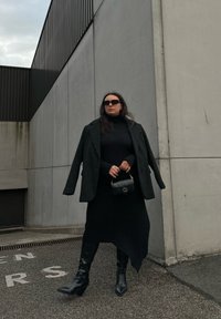 Črno oversized blazer nad prilegajočim črnim puloverjem s turtleneck ovratnikom, v kombinaciji z dolgo krilo in črnimi čevlji na peto. Drži majhno črno torbico. Betonska kulisa.