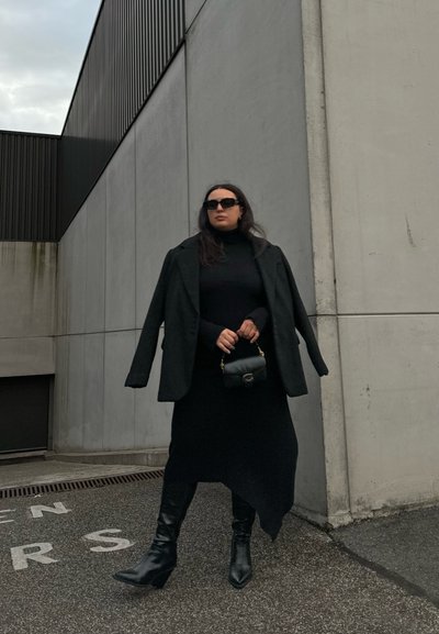 Blazer negro oversize sobre un ajustado cuello de tortuga negro, combinado con una falda larga y botas de tacón negras. Sosteniendo un pequeño bolso negro. Fondo de concreto.
